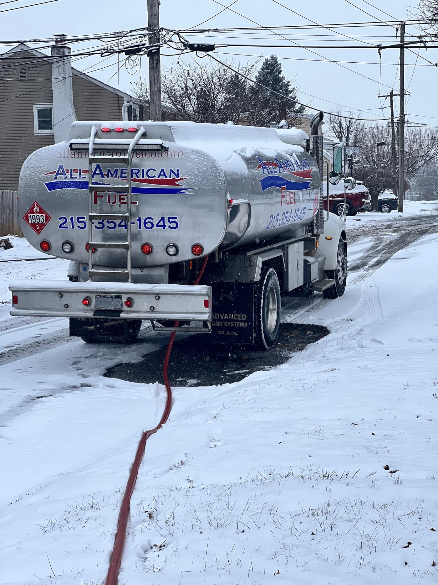 The rear of an All American Fuel tanker with the delivery hose running through the snow to a home.