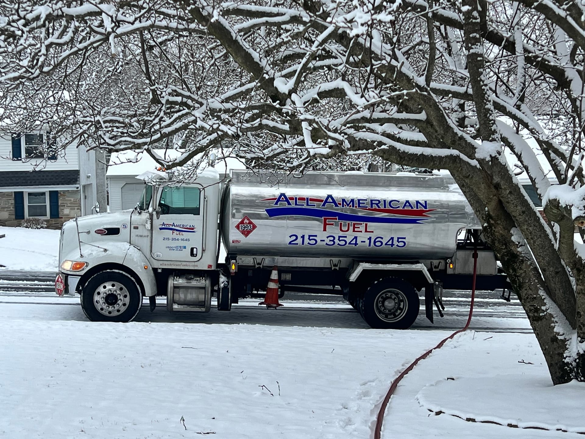 An All American Fuel tanker truck delivering heating oil on a snow-covered residential street.