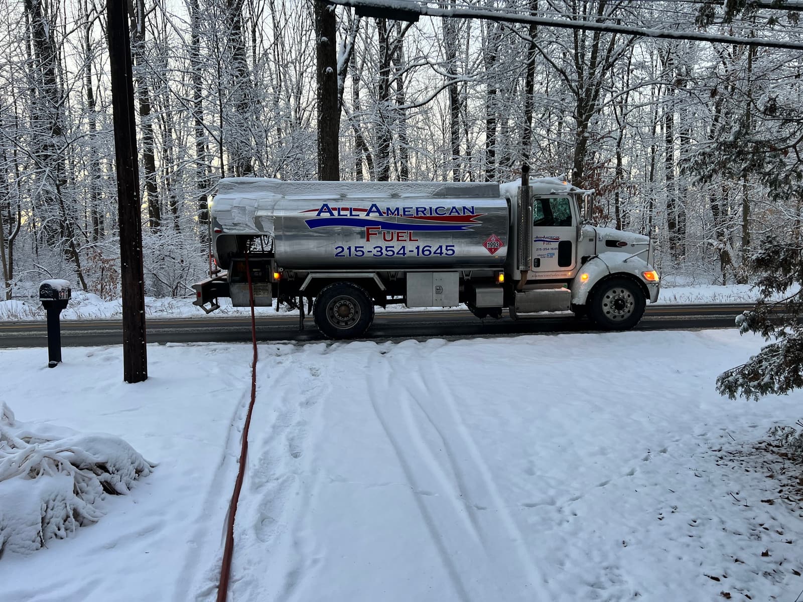 An All American Fuel truck parked on a snowy road with the delivery hose running across a frozen yard to a customer's home.